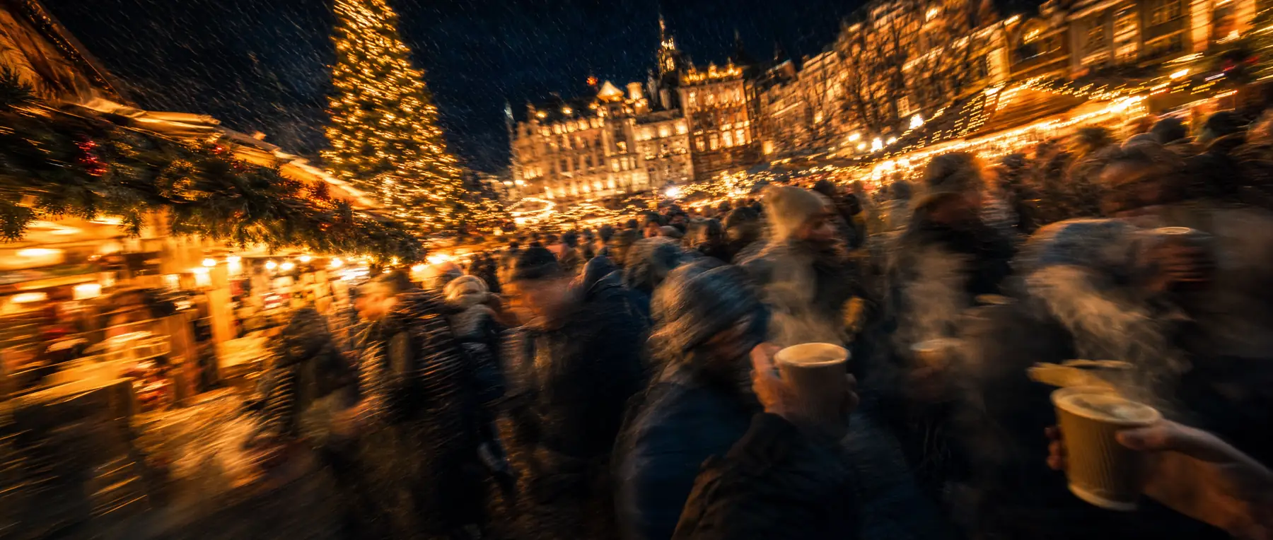 Weihnachtsmarkt bei Nacht in starker Bewegungsunschärfe, Dampf über Punschbechern, hunderte kleine warme Lichter zu Streifen verwischt