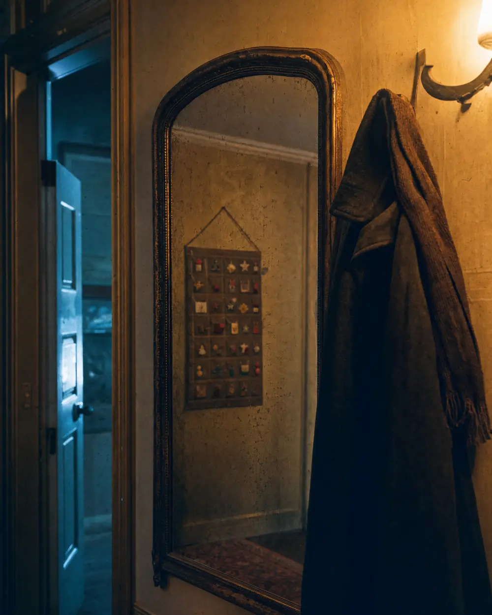 Alter Flurspiegel in einer dämmrigen Wohnung, daneben hängen Wintermantel und Schal, im Spiegel ist ein Adventskalender an der gegenüberliegenden Wand zu sehen